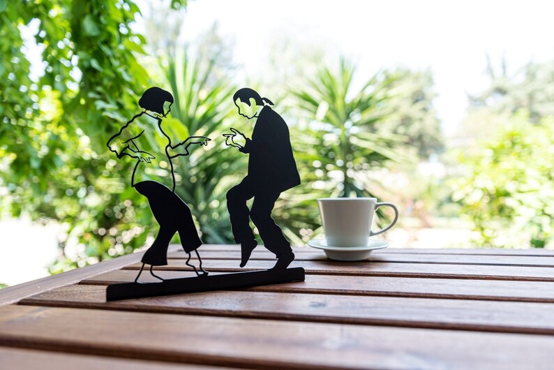 May include: A black metal silhouette sculpture depicting two figures dancing, mounted on a rectangular base. Beside the sculpture sits a white ceramic coffee cup and saucer. The figures are captured in a dynamic pose with arms extended.