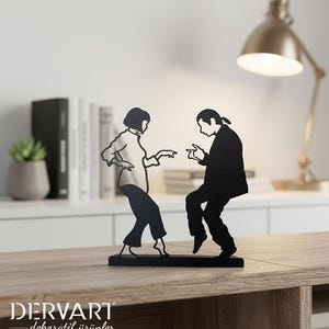 May include: A black metal silhouette sculpture depicting two figures in a dancing pose, featuring a person with a bob haircut and a person with a ponytail, both captured in a mid-motion stance, mounted on a rectangular black metal base.