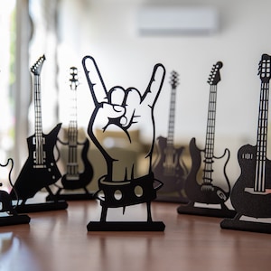 May include: A set of seven black metal guitar silhouettes, including a hand making the rock and roll hand gesture. The silhouettes are on a wooden surface.