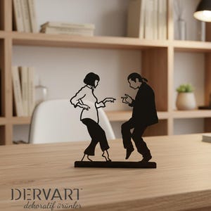 May include: A black metal silhouette sculpture depicting two figures in a dancing pose, featuring a person with a bob haircut and a person with long hair tied back, both wearing shirts and trousers, mounted on a rectangular base with the text DERVART.