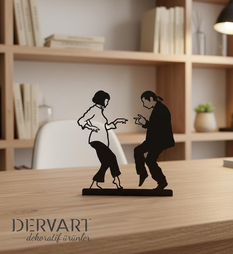 May include: A black metal silhouette sculpture depicting two figures in a dancing pose, featuring a person with a bob haircut and a person with long hair tied back, both wearing shirts and trousers, mounted on a rectangular base with the text DERVART.