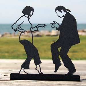 May include: A black metal silhouette sculpture depicting two figures dancing. The figures are shown in a side-by-side pose with arms extended and fingers pointing, mounted on a solid black rectangular base. The design features clean cutouts.
