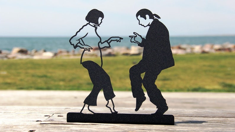 May include: A black metal silhouette sculpture depicting two figures dancing. The figures are shown in a side-by-side pose with arms extended and fingers pointing, mounted on a solid black rectangular base. The design features clean cutouts.