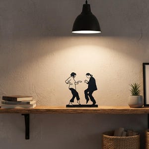 May include: A black metal silhouette sculpture depicting two figures in a dancing pose, featuring a person with a bob haircut and a person with long hair, both dressed in formal attire, standing on a flat base as a decorative tabletop ornament.