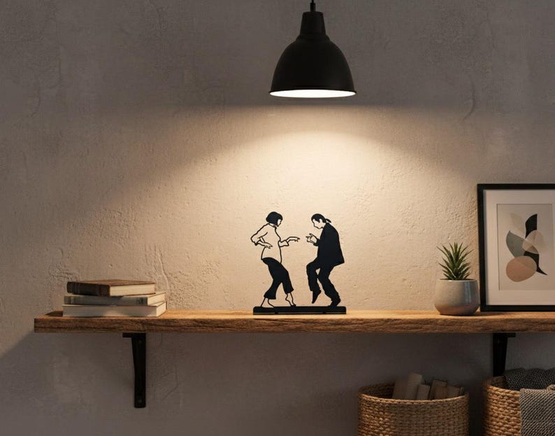 May include: A black metal silhouette sculpture depicting two figures in a dancing pose, featuring a person with a bob haircut and a person with long hair, both dressed in formal attire, standing on a flat base as a decorative tabletop ornament.