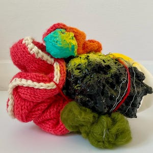 Pode incluir: Escultura abstrata com uma combinação de texturas e cores. A peça inclui uma seção de malha vermelha brilhante com acabamento branco, uma área texturizada preta e detalhes em verde, laranja, amarelo e turquesa. A escultura está sobre um fundo branco.