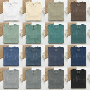 K&ouml;nnte beinhalten: Eine Sammlung gefalteter T-Shirts in verschiedenen Farben, darunter Wei&szlig;, Beige, Olivgr&uuml;n, Braun, Hellgr&uuml;n, T&uuml;rkis, Blau, Marine, Grau und Schwarz. Jedes Hemd ist ordentlich auf einer wei&szlig;en Oberfl&auml;che mit dekorativen Elementen angeordnet.