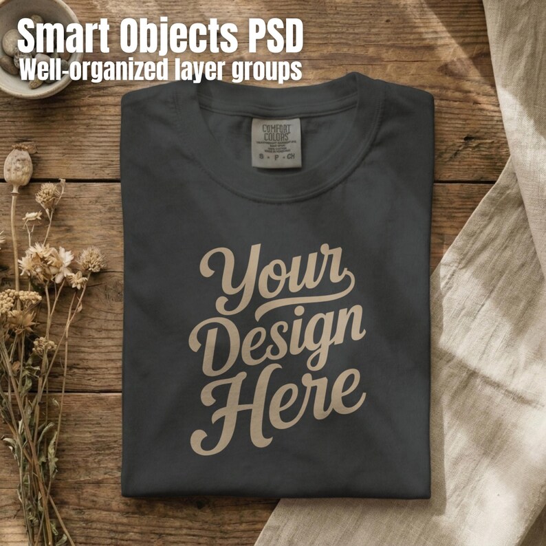 May include: A dark gray t-shirt featuring the text Your Design Here in a light tan script font. The shirt has a visible neck label with the text Comfort Colors S P CH. The design is centered on the chest of the folded garment.