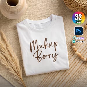 Könnte beinhalten: Ein gefaltetes weißes T-Shirt mit dem braunen Schriftzug "Mockup Berry". Das Shirt liegt auf einer beigen Oberfläche, mit einer dekorativen Vase und Holzperlen. Eine Grafik zeigt 32 Farboptionen und die Kompatibilität mit Photoshop, Canva und Photopea an.