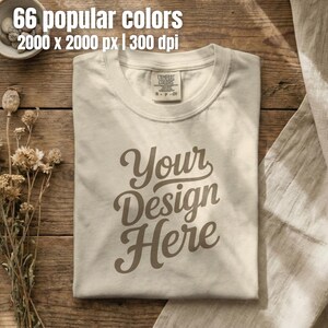 May include: A cream-colored short-sleeve t-shirt featuring the text Your Design Here in a dark brown script font. The shirt has a visible inner neck label that reads Comfort Colors, S P CH, and is made of 100% cotton fabric.