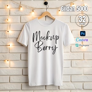 Può includere: Mockup di una t-shirt bianca con il testo "Mockup Berry" stampato sul davanti. La t-shirt è appesa a un appendiabiti di legno contro un muro di mattoni bianchi con luci a catena.