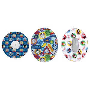 Op de afbeelding: Drie ronde plakstrips met Thomas de Tank Engine en zijn vrienden. De plakstrips zijn ontworpen om medische apparaten te bedekken. De plakstrips zijn blauw, groen en wit met verschillende afbeeldingen van Thomas en zijn vrienden.