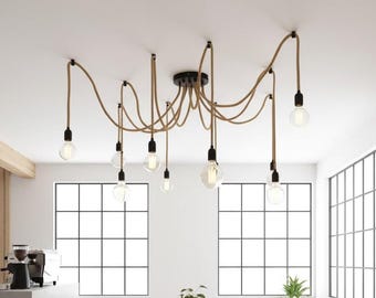 May include: A modern, multi-light pendant lamp with a black ceiling mount and tan cords. The cords extend to hold Edison-style light bulbs. The fixture is in a white room with large windows and a coffee machine.