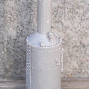 Könnte beinhalten: Ein graues Plastikmodell eines Dampfkessels mit einem hohen Schornstein und Nieten auf der Oberfläche. Das Modell hat eine kleine Tür an der Seite und ein Rohr oben.