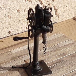 Pode incluir: Saca-rolhas antigo de metal preto com manivela e rodas decorativas. O saca-rolhas tem um parafuso sem fim longo e espiralado e é montado numa base quadrada. O design lembra máquinas vintage.