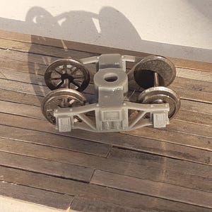 May include: A grey model train bogie with four wheels. The wheels are silver and black. The bogie is made of plastic and has a detailed design. It rests on a wooden surface.