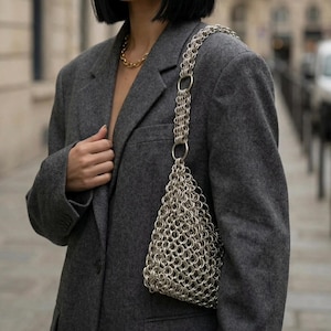May include: A silver chainmail bag with a triangular design and a chain strap. The bag is worn over the shoulder of a person in a grey blazer and a gold chain necklace. The background is blurred.
