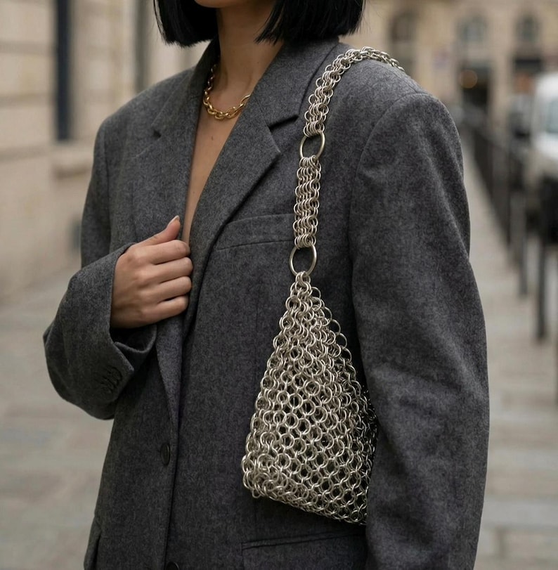 May include: A silver chainmail bag with a triangular design and a chain strap. The bag is worn over the shoulder of a person in a grey blazer and a gold chain necklace. The background is blurred.