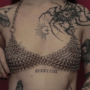 Może przedstawiać: Srebrzysty top typu halter wykonany z zazębiających się metalowych kółek. Odzież ma trójkątny krój z cienkim łańcuszkiem, który owija się wokół szyi, tworząc metaliczną, siatkową teksturę na klatce piersiowej.