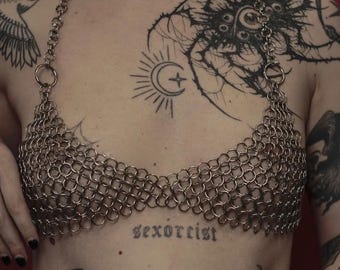 May include: A silver-toned chainmail halter top constructed from interlocking metal rings. The garment features a triangular bodice design with a thin chain strap that loops around the neck, creating a metallic, mesh-like texture across the chest area.