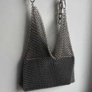 Könnte beinhalten: Eine silberfarbene Kettenbeuteltasche mit V-förmigem Design. Die Tasche wird an einer Kette und einem Metallverschluss aufgehängt. Die Kettenrüstung besteht aus kleinen, ineinandergreifenden Metallringen, die eine strukturierte Oberfläche bilden. Die Tasche ist leer und hängt an einer weißen Wand.