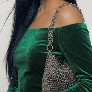 May include: A dark green velvet off-the-shoulder top with long sleeves. A silver chainmail bag with a chain strap hangs from the shoulder. The bag is made of small, interlocking metal rings.