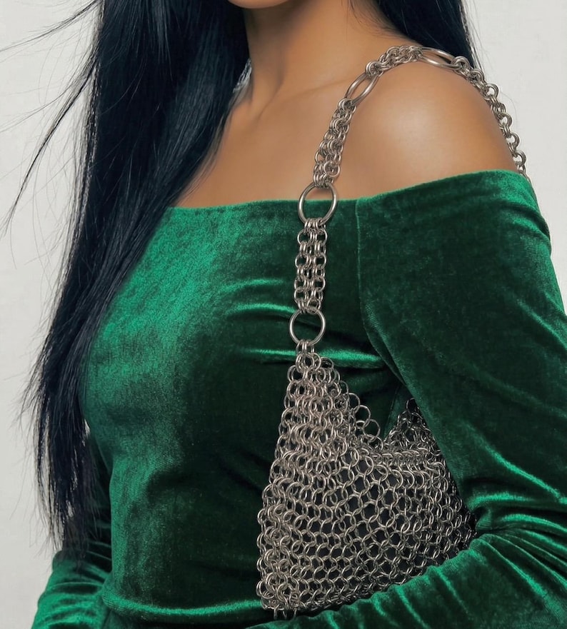 May include: A dark green velvet off-the-shoulder top with long sleeves. A silver chainmail bag with a chain strap hangs from the shoulder. The bag is made of small, interlocking metal rings.