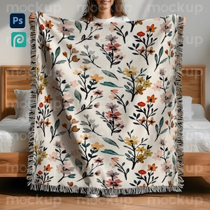 May include: A cream-coloured woven throw blanket with a floral pattern in shades of orange, yellow, pink, and green. The blanket has a fringed edge and is displayed against a wooden bed frame. The floral design features various flower and leaf shapes.