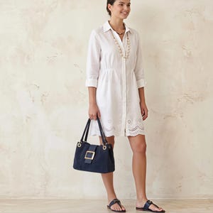 May include: A white button-down shirt dress with three-quarter sleeves and a scalloped eyelet hem, paired with a navy blue handbag featuring a large gold-tone buckle and gold hardware, and dark navy blue thong sandals with gold-tone accents.