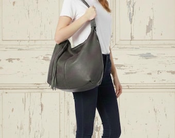 May include: A gray leather hobo bag with a braided shoulder strap and a tassel detail. The bag is carried over the shoulder. The background is a white door with peeling paint.