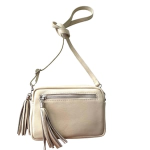 May include: A small, off-white leather crossbody bag with a zipped pocket and two tassels. The bag has a long adjustable strap.