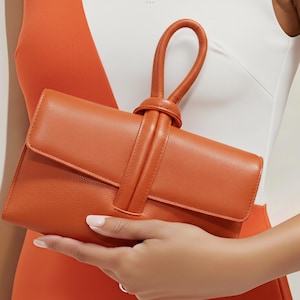 May include: A burnt orange leather clutch with a flap closure and a looped handle. The clutch is rectangular and features a textured surface. The handle is attached to the top of the bag with a knotted detail.
