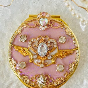 Può includere: Uno specchio compatto rosa con una cornice dorata e un design intricato. Lo specchio è decorato con strass, perle e una corona dorata con ali.