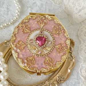 Könnte beinhalten: Ein dekorativer, herzförmiger Kompaktspiegel mit rosa Glitzerhintergrund und goldenen Filigrandetails. In der Mitte befindet sich ein rosa herzförmiger Edelstein, umgeben von kleineren Edelsteinen. Eine Perlenkette ist sichtbar.