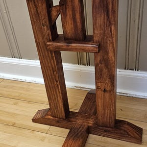 May include: A handcrafted wooden stand with a dark stain. The stand features a unique design with angled legs and a notched top. The wood grain is visible, adding to its rustic charm. The stand is likely for display or decorative purposes.