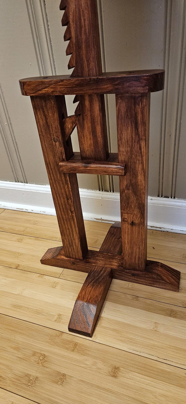 May include: A handcrafted wooden stand with a dark stain. The stand features a unique design with angled legs and a notched top. The wood grain is visible, adding to its rustic charm. The stand is likely for display or decorative purposes.