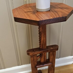 May include: A wooden plant stand with a dark stain. The octagonal tabletop holds a white ceramic pot with a green plant. The stand has a carved central support and a solid base. The stand is approximately 76 cm tall.