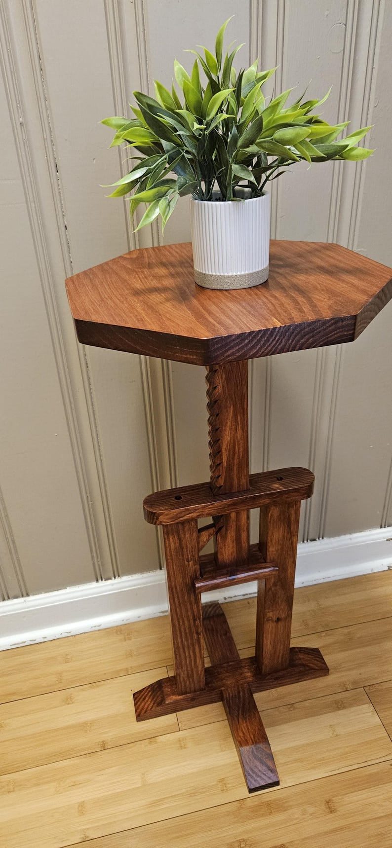 May include: A wooden plant stand with a dark stain. The octagonal tabletop holds a white ceramic pot with a green plant. The stand has a carved central support and a solid base. The stand is approximately 76 cm tall.