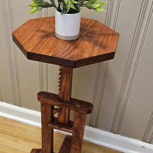 May include: A wooden plant stand with a hexagonal top and adjustable height. The stand is stained a rich brown and supports a small white pot with a green plant. The design features a unique, adjustable mechanism. The stand's height is adjustable.