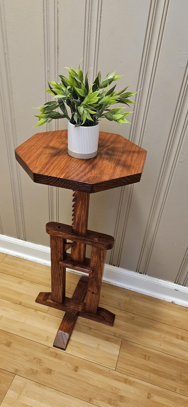 May include: A wooden plant stand with a hexagonal top and adjustable height. The stand is stained a rich brown and supports a small white pot with a green plant. The design features a unique, adjustable mechanism. The stand's height is adjustable.