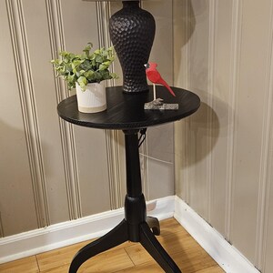 May include: A black accent table with a round top, featuring a decorative lamp with a brown lampshade, a small potted plant, and a red cardinal figurine. The table has a three-legged base.