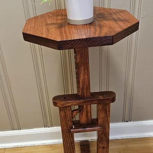 May include: A handcrafted wooden plant stand with a hexagonal top and a dark stain. The stand features a decorative support structure and holds a small white pot with a green plant. The stand is approximately 30 inches tall.