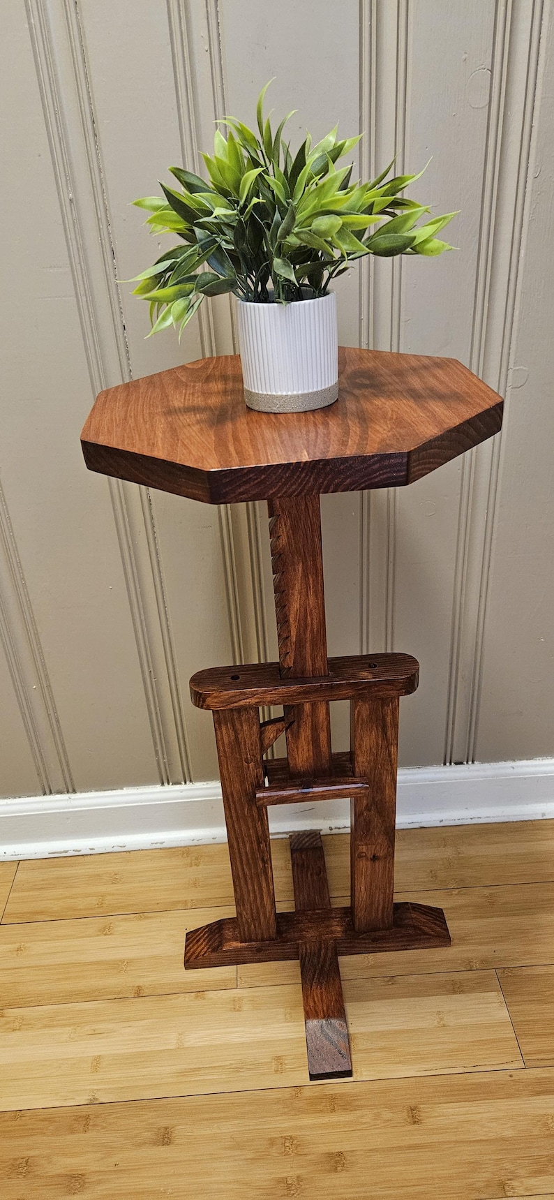 May include: A handcrafted wooden plant stand with a hexagonal top and a dark stain. The stand features a decorative support structure and holds a small white pot with a green plant. The stand is approximately 76 cm tall.
