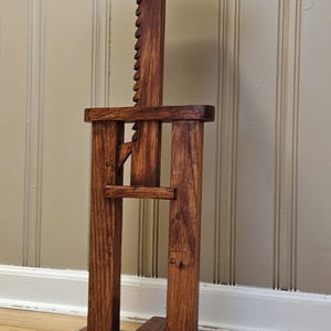 May include: A handcrafted wooden plant stand with a dark stain. The stand features a multi-sided top, a central support column with a notched adjustment, and a cross-shaped base. The stand holds a green plant.