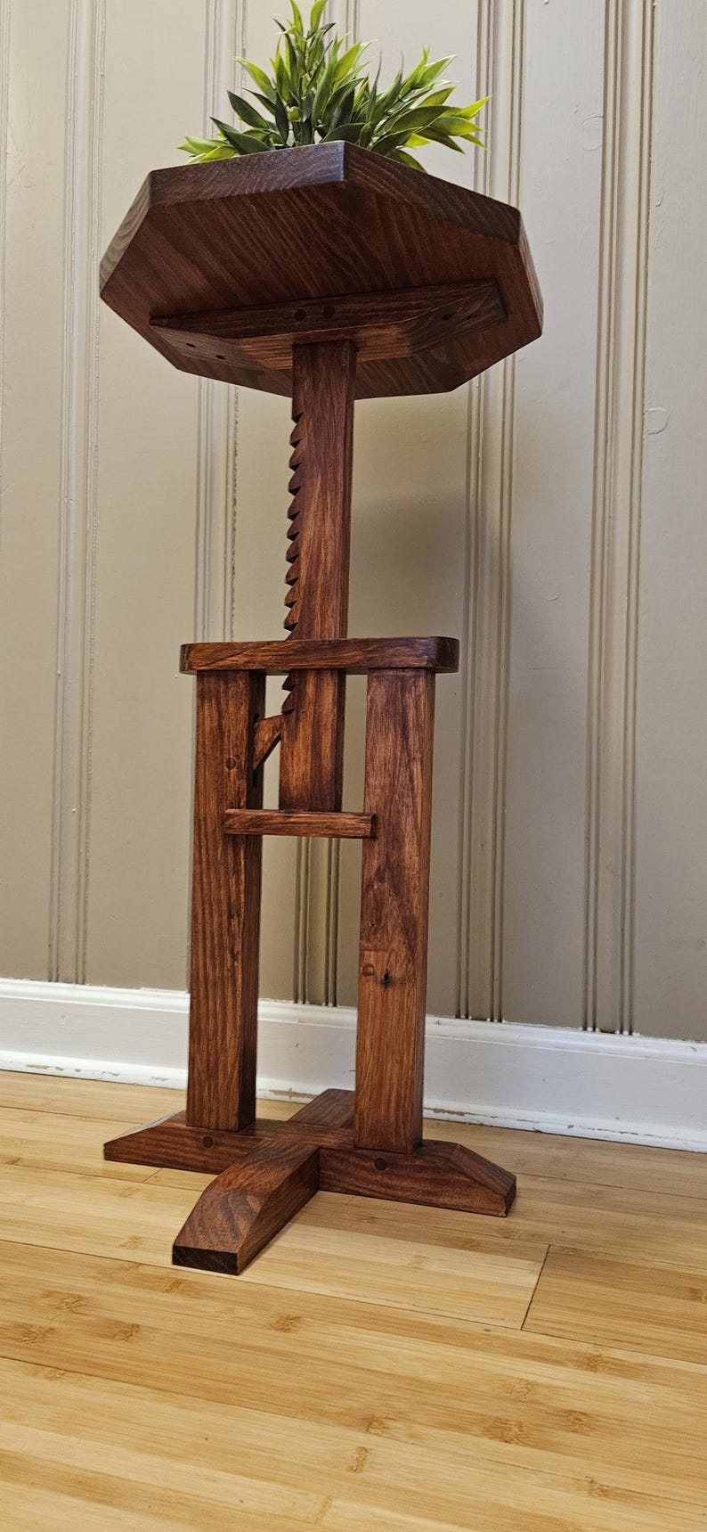 May include: A handcrafted wooden plant stand with a dark stain. The stand features a multi-sided top, a central support column with a notched adjustment, and a cross-shaped base. The stand holds a green plant.