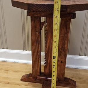 May include: A handcrafted wooden side table with a dark stain. The table has a unique design with a geometric top and decorative cutouts. The table is approximately 58 cm tall. A white vase with greenery sits on top.
