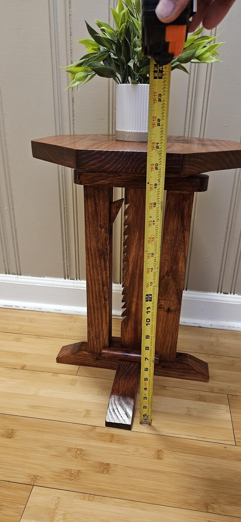 May include: A handcrafted wooden side table with a dark stain. The table has a unique design with a geometric top and decorative cutouts. The table is approximately 58 cm tall. A white vase with greenery sits on top.