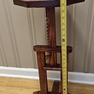 May include: A handcrafted wooden plant stand with a dark stain. The stand has an octagonal top and a unique, geometric base. A white and tan ceramic pot with greenery sits on top. The stand is approximately 91 cm tall.