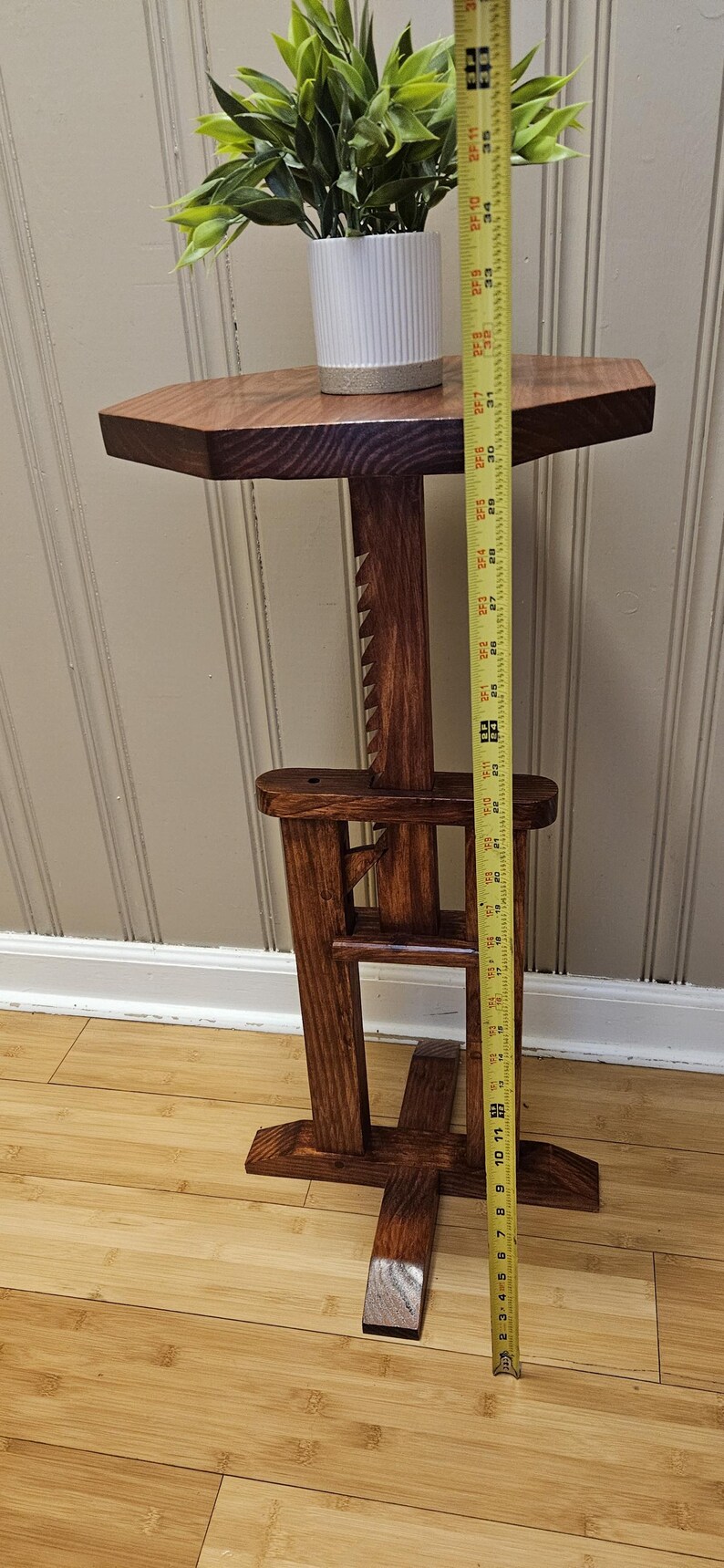 May include: A handcrafted wooden plant stand with a dark stain. The stand has an octagonal top and a unique, geometric base. A white and tan ceramic pot with greenery sits on top. The stand is approximately 91 cm tall.