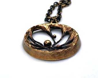 Puede incluir: Un collar con un colgante de bronce de diseño circular que presenta una rama de árbol estilizada con pequeñas esferas. El colgante está unido a una cadena.